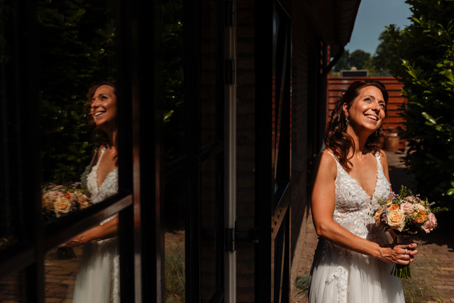 real bride sanneke – romantische A-lijn trouwjurk – bruid lacht terwijl ze buiten staat met een pastelboeket; haar reflectie is prachtig zichtbaar in het raam naast haar